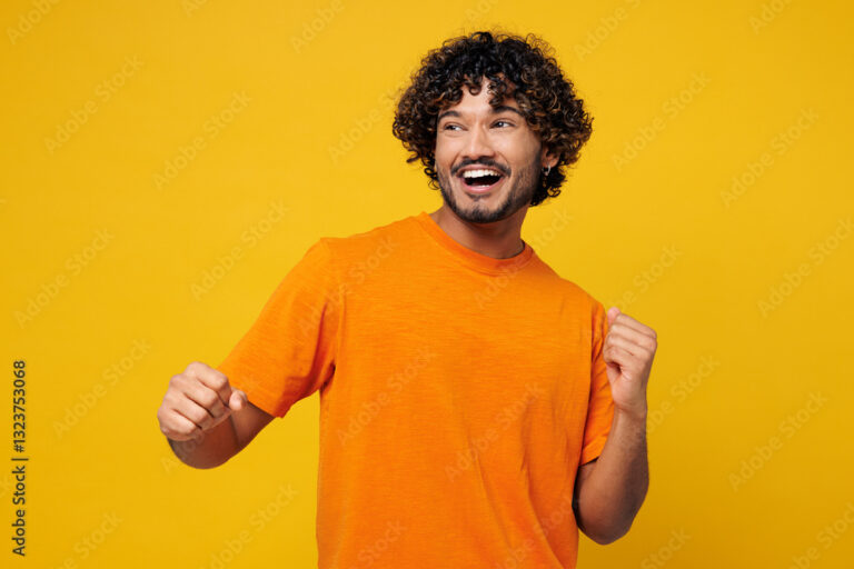 Young smiling fun happy Indian man wear orange t-shirt casual clothes doing winner gesture celebrate clenching fists dancing look aside on area isolated on plain yellow background. Lifestyle concept.